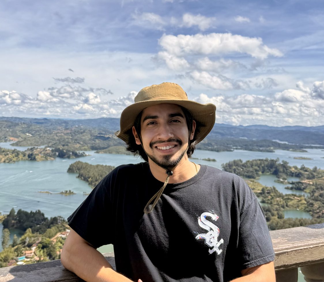 Jorge Cazares, in a bucket hat and black t-shirt, smiling to camera from a balcony above a bright blue reservoir dotted with green islands.